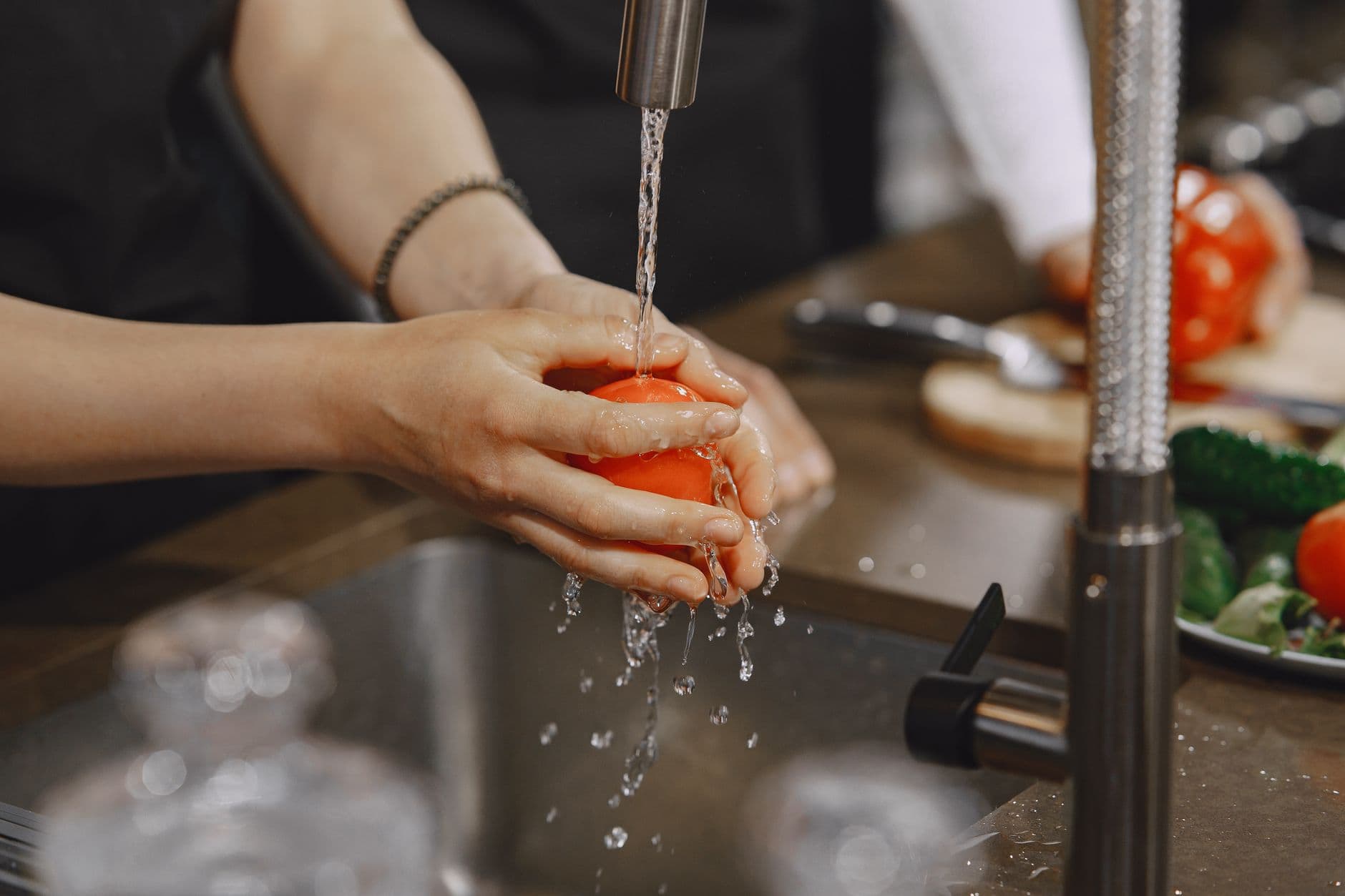 Hands rinsing a tomato under running water at a kitchen faucet